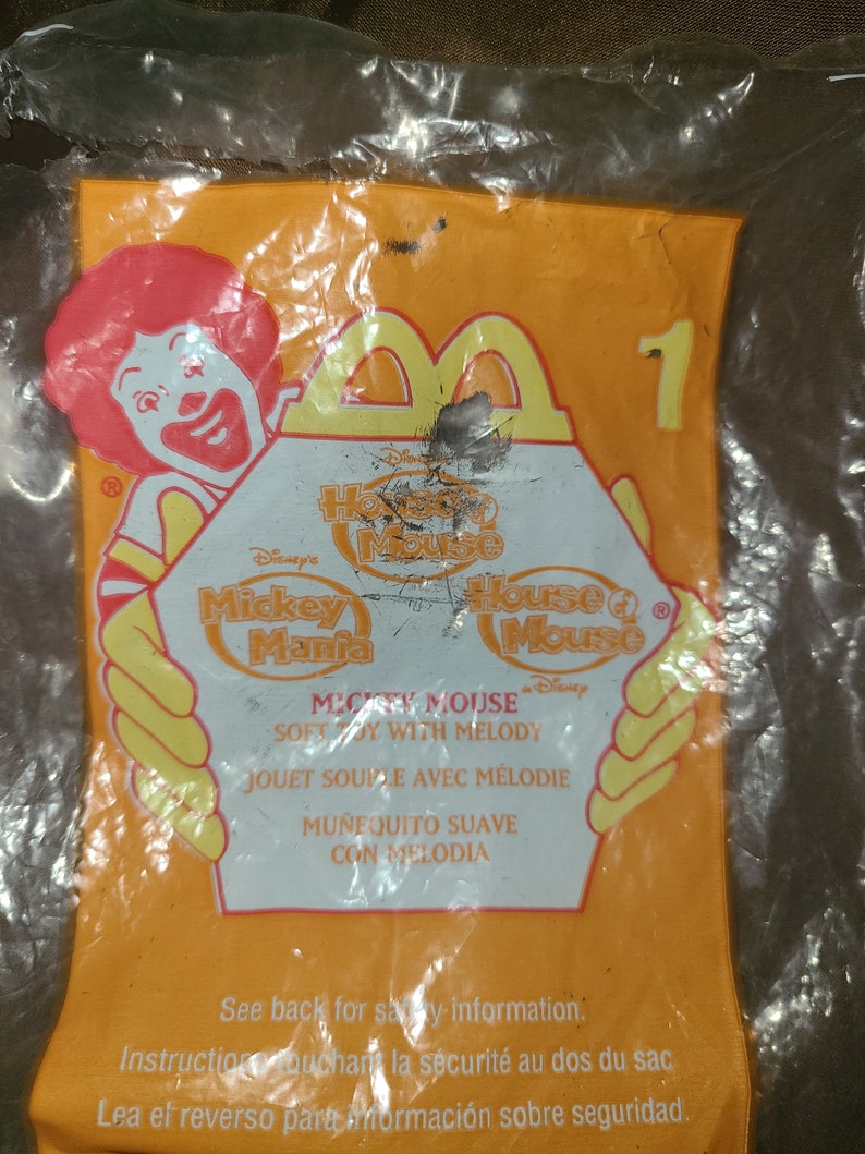 May include: A yellow plastic bag with a Mickey Mouse Mania toy inside. The bag features a red and yellow Mickey Mouse character and the text "Mickey Mouse Mania" and "House of Mouse".