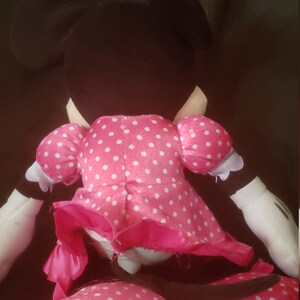 Pu&ograve; includere: Un giocattolo di peluche rosa Minnie Mouse con pois bianchi. Il giocattolo &egrave; seduto su una superficie scura.