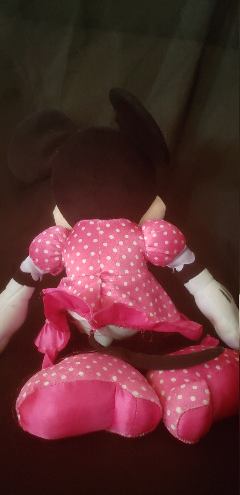Pu&ograve; includere: Un giocattolo di peluche rosa Minnie Mouse con pois bianchi. Il giocattolo &egrave; seduto su una superficie scura.