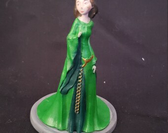 May include: A green and gold painted figurine of a woman wearing a long green dress and a gold chain belt. The figurine is standing on a round gray base.