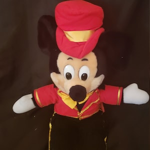 May include: A plush Mickey Mouse toy wearing a red and gold conductor's hat and a red jacket with gold trim. The toy has black pants and yellow shoes.