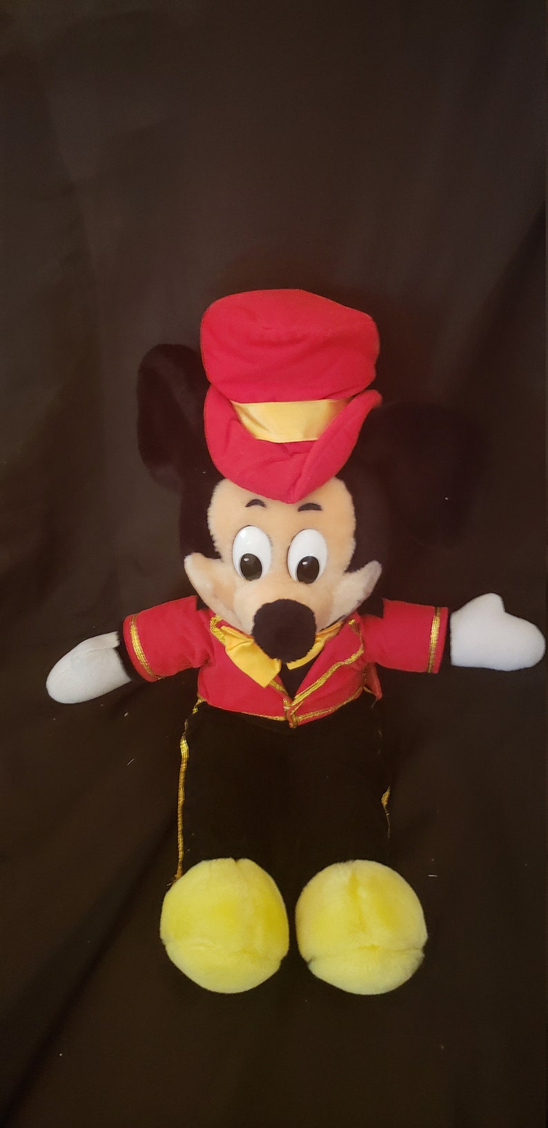 May include: A plush Mickey Mouse toy wearing a red and gold conductor's hat and a red jacket with gold trim. The toy has black pants and yellow shoes.