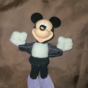 May include: A plush Mickey Mouse toy wearing a black suit with a white shirt and purple shoes.