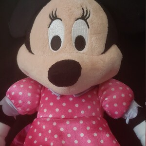 Pu&ograve; includere: Un giocattolo di peluche rosa con pois bianchi, che rappresenta un topo dei cartoni animati con occhi grandi e ciglia nere. Il giocattolo indossa un vestito rosa con un colletto bianco e un orlo arricciato.