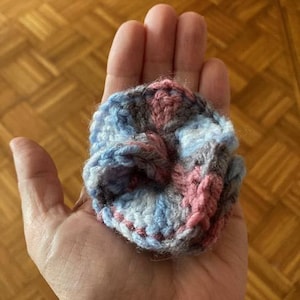 May include: A handmade crochet flower in shades of blue, pink, and gray. The flower is held in a hand, showcasing the intricate stitch work and color variations. The craft item is a small, decorative piece.