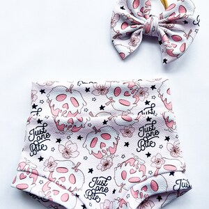 May include: A pair of high-waisted bloomers and a matching hair bow featuring a white background with a repeating pattern of pink skull-shaped apples, small flowers, black stars, and the black text "Just one Bite" printed across the fabric.