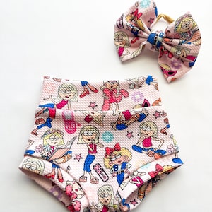 May include: A baby clothing set featuring a pink high-waisted short and a matching bow. Both items are covered in a cartoon print of a blonde girl in various poses. The bow is attached to a yellow elastic band.