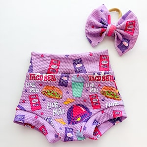 May include: A set of baby shorts and a matching bow. The shorts and bow are a light purple color with a repeating pattern of Taco Bell-themed graphics, including the Taco Bell logo, tacos, and sauce packets.
