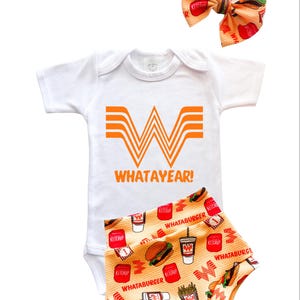 May include: A white baby onesie with an orange logo and the text "WHATAYEAR!". The set includes matching orange and white patterned shorts and a bow. The bow and shorts feature images of burgers, fries, and ketchup.