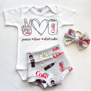May include: A white baby bodysuit, grey shorts, and a matching bow. The bodysuit features a peace sign, heart, and Diet Coke can graphic with the words "peace love diet coke." The shorts and bow have a Diet Coke and daisy print.