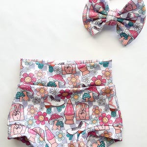 May include: A white and purple checkered baby bloomer with a floral and retro design featuring mushrooms, peace signs, and roller skates. A matching headband with a large bow is also included.