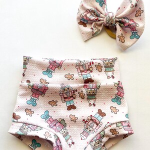 Puede incluir: Shorts de bebé rosas y un lazo a juego con un estampado repetido de hombrecitos de jengibre, cascanueces y cupcakes. Los shorts tienen cintura alta y borde con volantes. El lazo está atado con una banda elástica amarilla.