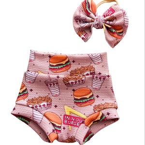 May include: A pink fabric set consisting of high-waisted shorts and a matching hair bow. The fabric features a repeating pattern of hamburgers, french fries, and drink cups, with a yellow sign displaying the text IN-N-OUT BURGER.