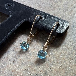 May include: A pair of gold-colored drop earrings with round, light blue gemstones. The earrings are displayed against a textured, neutral-toned surface and a black object.