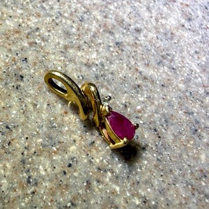 May include: A gold pendant with a teardrop-shaped, deep red gemstone. The pendant has a loop for a chain and a small, clear accent stone. The setting is a bright, polished gold.