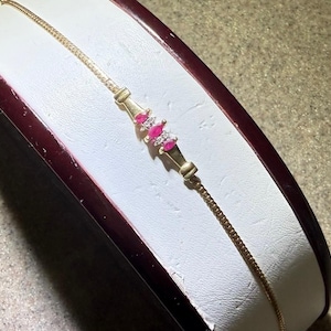 May include: A delicate gold chain bracelet featuring a central decorative element set with three marquise-cut red rubies and small clear accent stones. The bracelet has a fine link chain design and a polished gold finish throughout the metal structure.