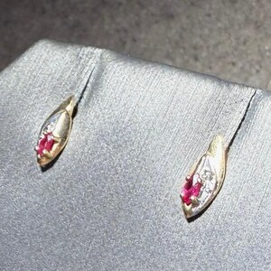 May include: A pair of gold-toned stud earrings. Each earring features a teardrop shape with a cluster of small, bright red gemstones and a few small white gemstones. The earrings are displayed on a gray surface.