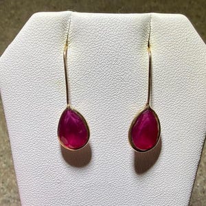 May include: A pair of gold-colored drop earrings with teardrop-shaped, deep pink gemstones. The gemstones are set in a gold-colored bezel and hang from long, slender gold-colored wires. The earrings are displayed on a white textured surface.