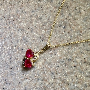 May include: A gold-colored necklace with a pendant featuring two heart-shaped, bright red gemstones. Small, clear stones are set around the red gems. The necklace chain is delicate and thin.