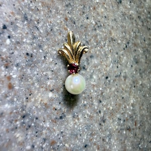 May include: A gold pendant featuring a stylized fleur-de-lis top, a small round red gemstone set in the center, and a single white spherical pearl dangling from the bottom. The metal has a polished finish and the design is symmetrical.