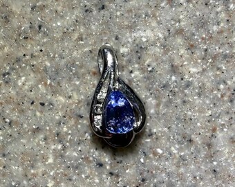 May include: A silver-toned pendant featuring a pear-shaped blue gemstone at the center, accented by three small clear round stones set along one side of the curved metal frame, which includes a bail at the top for attachment to a chain.