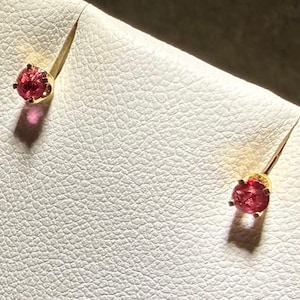 May include: A pair of stud earrings featuring round, faceted pink gemstones set in gold-toned metal prongs. Each earring is secured to a white textured display card, with the stones catching the light to create small, distinct shadows below.