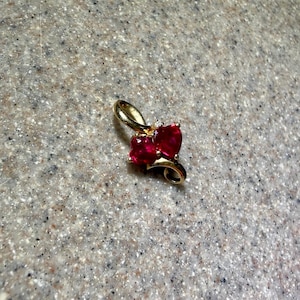 May include: A gold pendant featuring two heart-shaped red gemstones arranged side-by-side, accented by a single small clear stone at the top, with a curved gold bail extending from the upper portion of the heart-shaped setting.