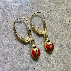 May include: A pair of gold-tone leverback earrings featuring dangling ladybug charms. Each charm is painted with a bright red body, black spots, and a black head. The metal has a polished finish, and the leverback mechanism is attached to the top.