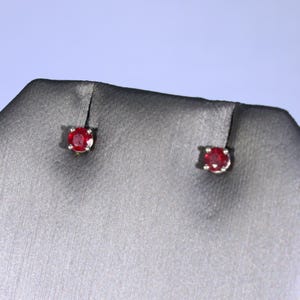 May include: A pair of stud earrings featuring round, faceted red gemstones set in a simple metal prong setting. The earrings are displayed on a dark gray textured surface, highlighting the vibrant red color and the circular cut of the stones.