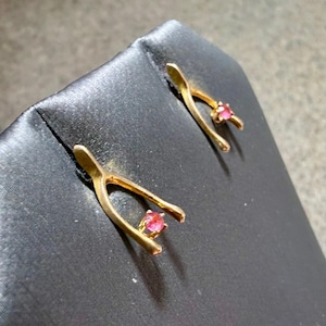 May include: A pair of gold-toned wishbone-shaped stud earrings. Each earring features a smooth, curved metal frame in a Y-shape, with a single small, round, vibrant pink gemstone set securely at the base where the two prongs meet.