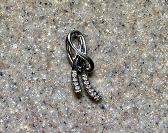 May include: A silver-toned pendant featuring a looped, ribbon-like design. The two lower ends of the pendant are embellished with a row of small, clear, sparkling stones that catch the light, creating a polished and reflective metallic appearance.