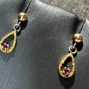 May include: A pair of gold-toned drop earrings. Each earring features a small, round stud connected to a textured, teardrop-shaped pendant. A small, red gemstone is set in the center of each pendant. The earrings are displayed against a dark background.
