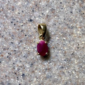 May include: A gold pendant with a vibrant, oval-shaped ruby gemstone. The ruby is a deep, rich red color, and the gold setting has a simple, elegant design. The pendant is suspended from a small loop at the top.