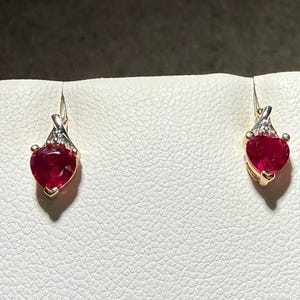 May include: A pair of gold earrings featuring heart-shaped red gemstones. Each earring has a small, triangular metal setting at the top accented with tiny clear stones, connecting to the vibrant red heart-shaped stone suspended below.