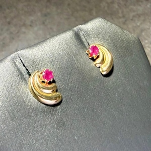 May include: A pair of gold-toned stud earrings, each featuring a bright pink gemstone. The earrings have a layered, curved design, and are displayed on a dark gray surface.