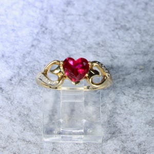 May include: A gold ring featuring a central heart-shaped red gemstone. The band has a decorative openwork design on both sides, accented with small clear stones embedded along the shoulders of the ring, creating a detailed and polished appearance.