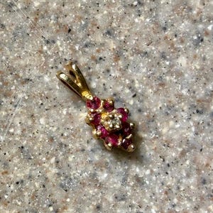 May include: A gold pendant featuring a cluster of small, round, deep red rubies arranged in a floral-like pattern, with a single clear, colorless diamond set in the center. The pendant includes a gold bail at the top for attaching to a chain.