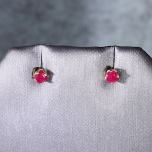 May include: A pair of stud earrings featuring round, faceted, deep pinkish-red gemstones set in gold-toned metal prongs. The stones are circular in shape and display a textured, natural appearance within the secure metal settings.