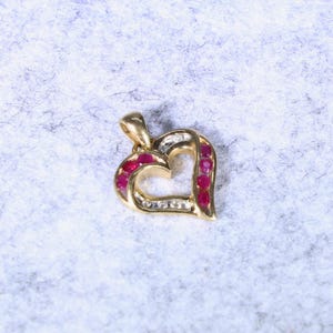 May include: A gold heart-shaped pendant featuring a series of small, round, vibrant red rubies set along the curved frame, accompanied by a row of tiny, clear, sparkling diamonds nestled within the polished gold metal structure.
