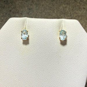 May include: A pair of gold-toned stud earrings, each featuring a light blue, oval-shaped gemstone. The earrings are set against a textured white surface.