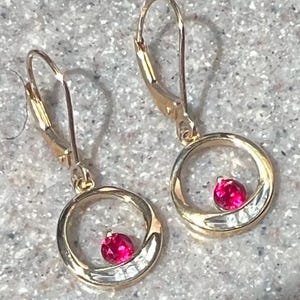May include: A pair of gold-toned earrings. Each earring features a circular design with a vibrant red gemstone and small, clear stones. The earrings have a lever-back closure.