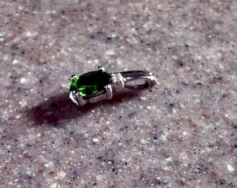 May include: A silver pendant with a vibrant green oval gemstone. The gemstone is set in a four-prong setting, and the pendant has a small loop for a chain. The pendant is displayed on a speckled surface.