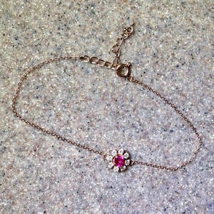 May include: A delicate rose gold bracelet featuring a floral design. The flower has a vibrant pink center stone surrounded by small, clear gemstones. The bracelet has an adjustable chain and a secure clasp.