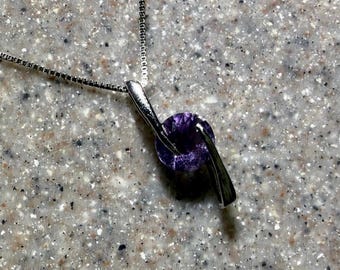 May include: A silver-toned pendant necklace featuring a round, faceted purple gemstone held by two curved, metallic prongs. The pendant is attached to a thin, square-link chain that extends upward from the top of the setting.