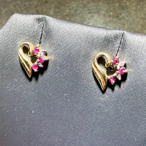 May include: A pair of gold heart-shaped stud earrings. Each earring features a smooth gold curved side and a cluster of three small round pink gemstones and one clear accent stone arranged along the opposite side of the heart frame.
