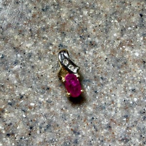 May include: A gold pendant with a ruby-colored oval gemstone and three small, clear stones. The pendant has a curved design with a gold base and a silver-colored top section. The gemstone is a vibrant, deep red color.