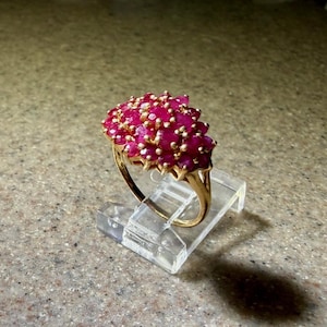 May include: A gold ring featuring a cluster of numerous small, round, vibrant pinkish-red gemstones set closely together in a dome-shaped arrangement. The band is thin and polished, supporting the dense, textured gemstone head of the ring.
