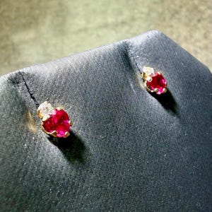 May include: A pair of gold stud earrings, each featuring a vibrant red gemstone and a cluster of small, clear stones. The earrings are displayed on a black surface, showcasing their sparkle and design.