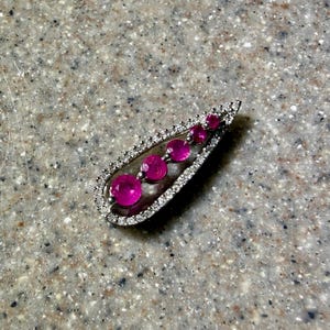 May include: A silver-toned pendant with a teardrop shape, featuring a row of five vibrant pink gemstones. The pendant's border is encrusted with small, clear crystals, creating a sparkling effect. The gemstones are round and evenly spaced.