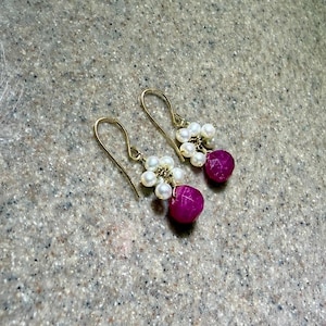 May include: A pair of drop earrings featuring a cluster of small white pearls arranged in a floral pattern, accented by a single faceted, deep pink gemstone bead suspended from a gold-toned wire hook. The design is symmetrical and delicate.